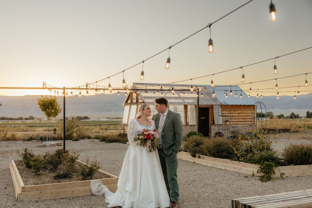Bride and groom embrace under string lights at sunset with rustic barn venue in the background.