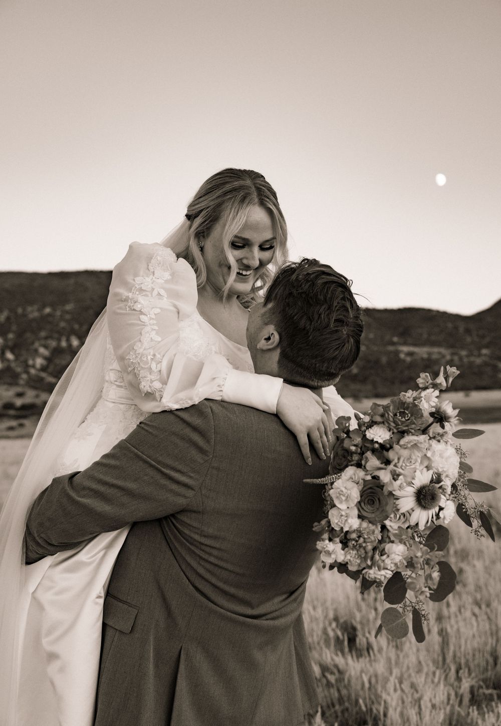 Black and white photo of groom lifting smiling bride in a field with mountains and moon in background.