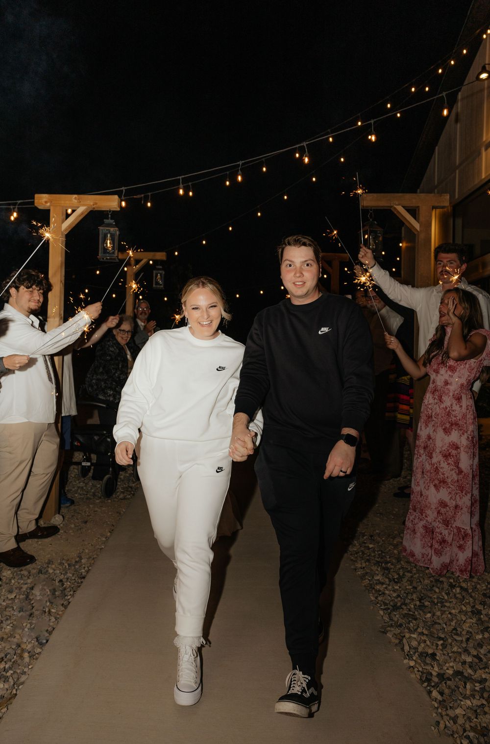 Newlyweds in matching Nike sweatsuits run through a sparkler send-off under string lights at night.