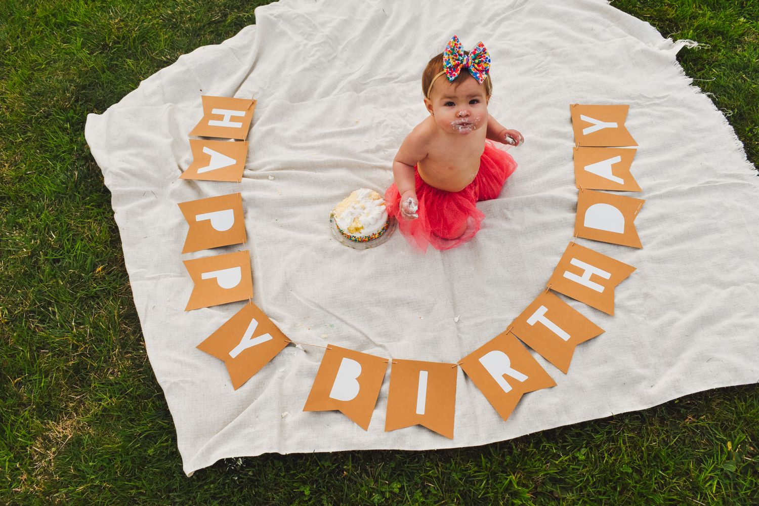 Baby in red tutu sits on white blanket with orange birthday lettering.