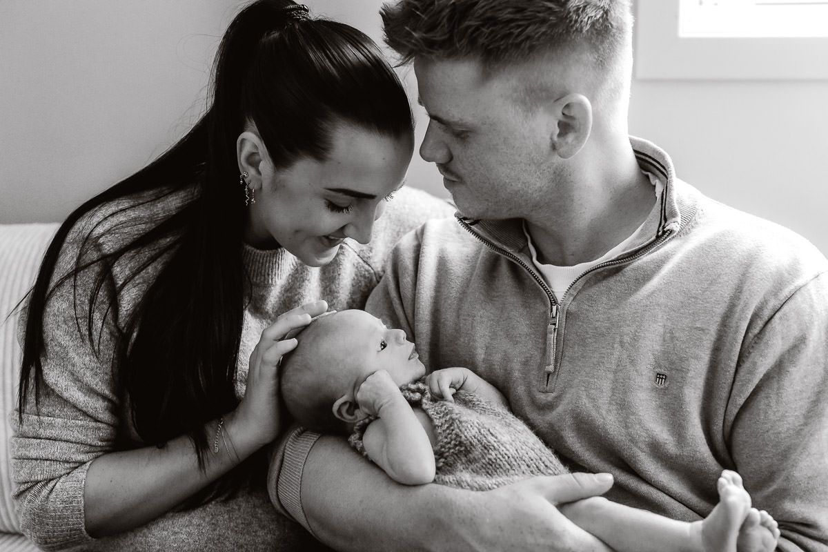 Svart-hvitt bilde av mamma, pappa og baby som sitter tett i sofa. Pappa ser kjærlig på mamma som tar på hodet til babyen.