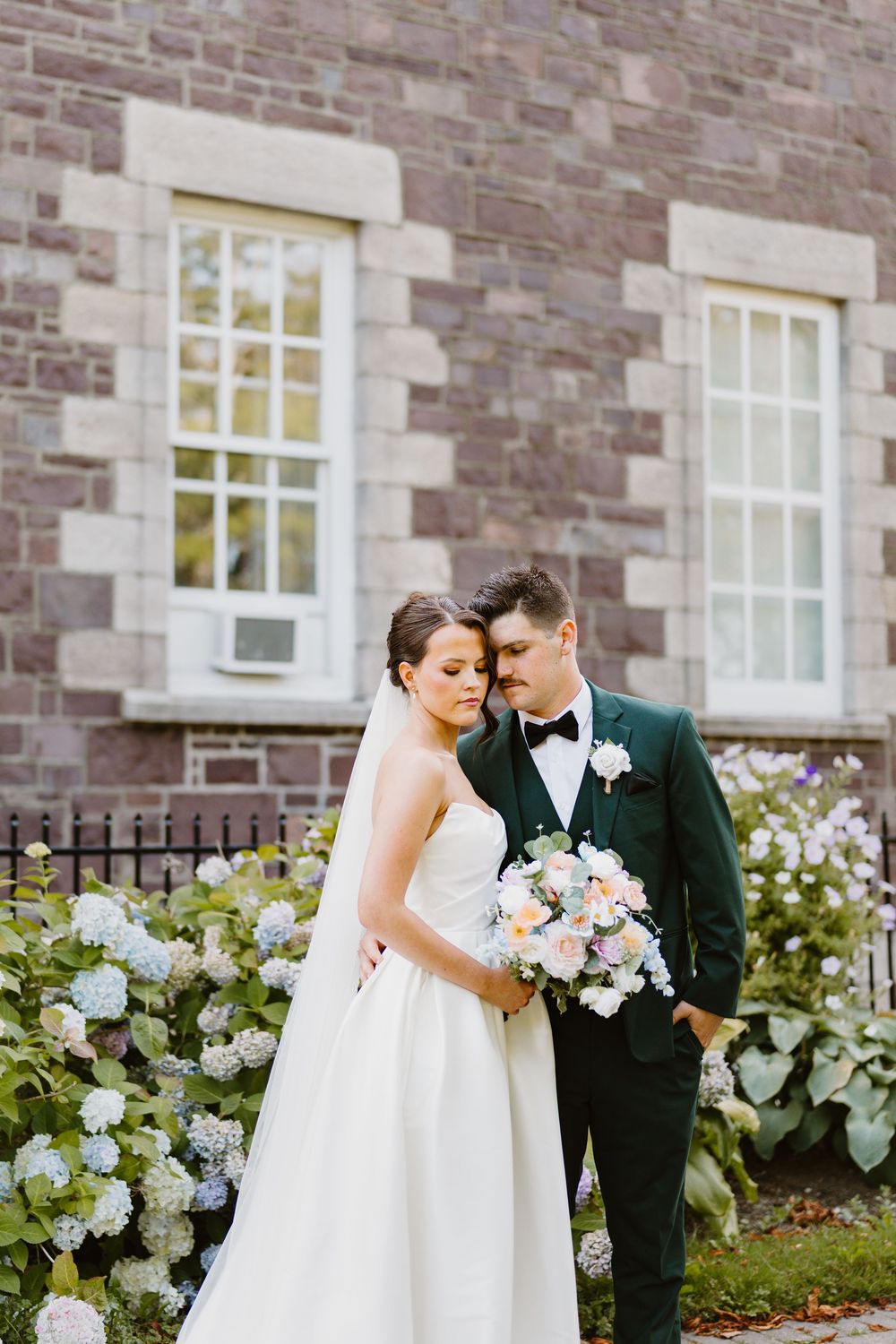 A romantic wedding couple shares a tender moment together in front of a stone building with white flowers blooming.