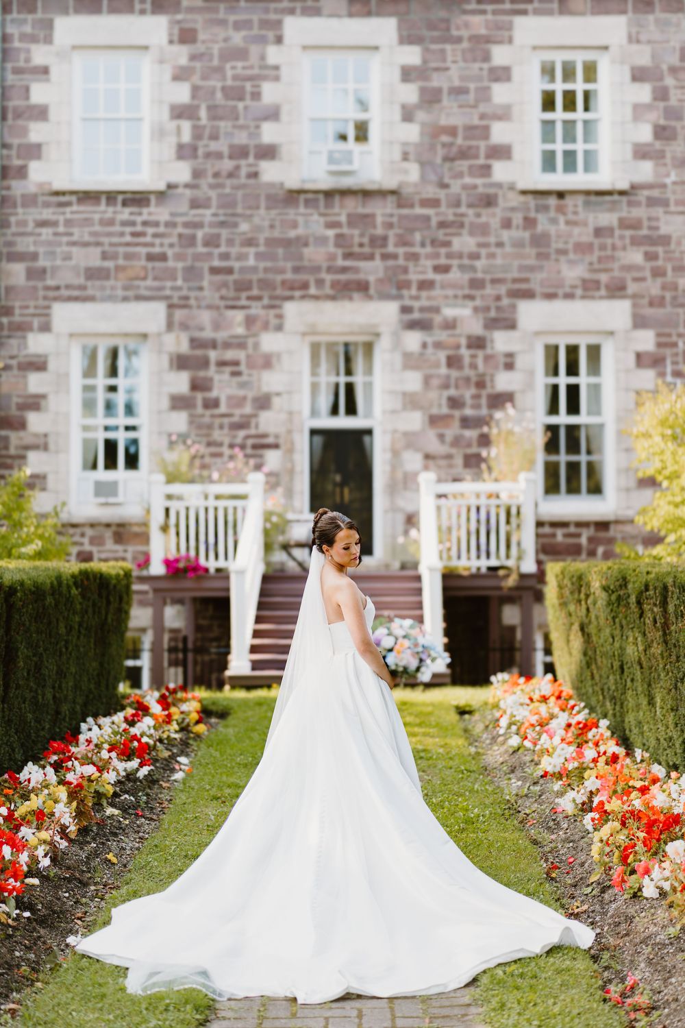 A wedding dress with long train photographed in front of a stone mansion with colorful garden flowers along the path.