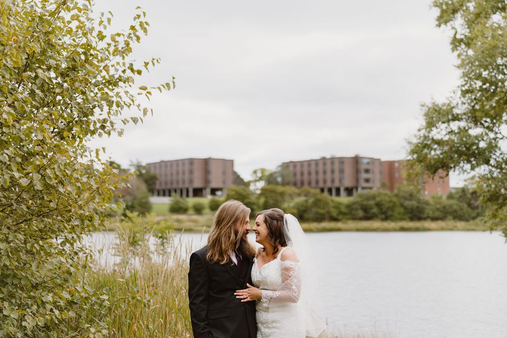 Two people embrace while standing near a waterfront with buildings and autumn trees in the background.