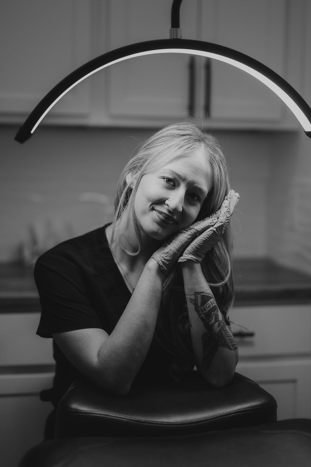 Black and white portrait of a smiling blonde woman in gloves resting her chin on hands under a curved light in a studio.