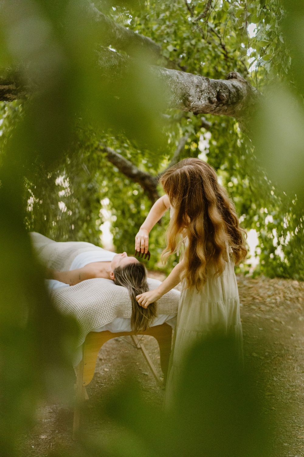 Woman in white dress lying peacefully on a large tree branch surrounded by green summer foliage.