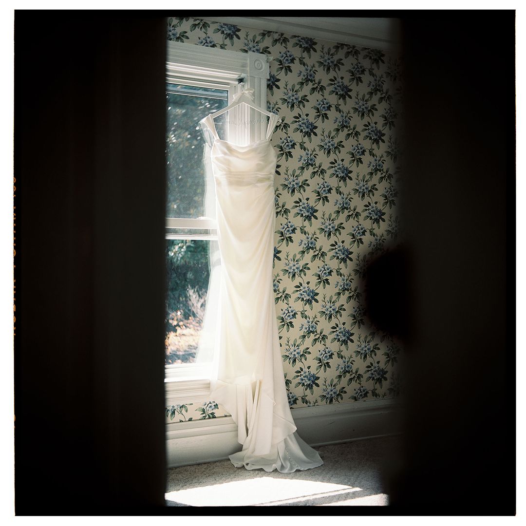 A white wedding dress hangs elegantly against blue floral wallpaper in natural window light.
