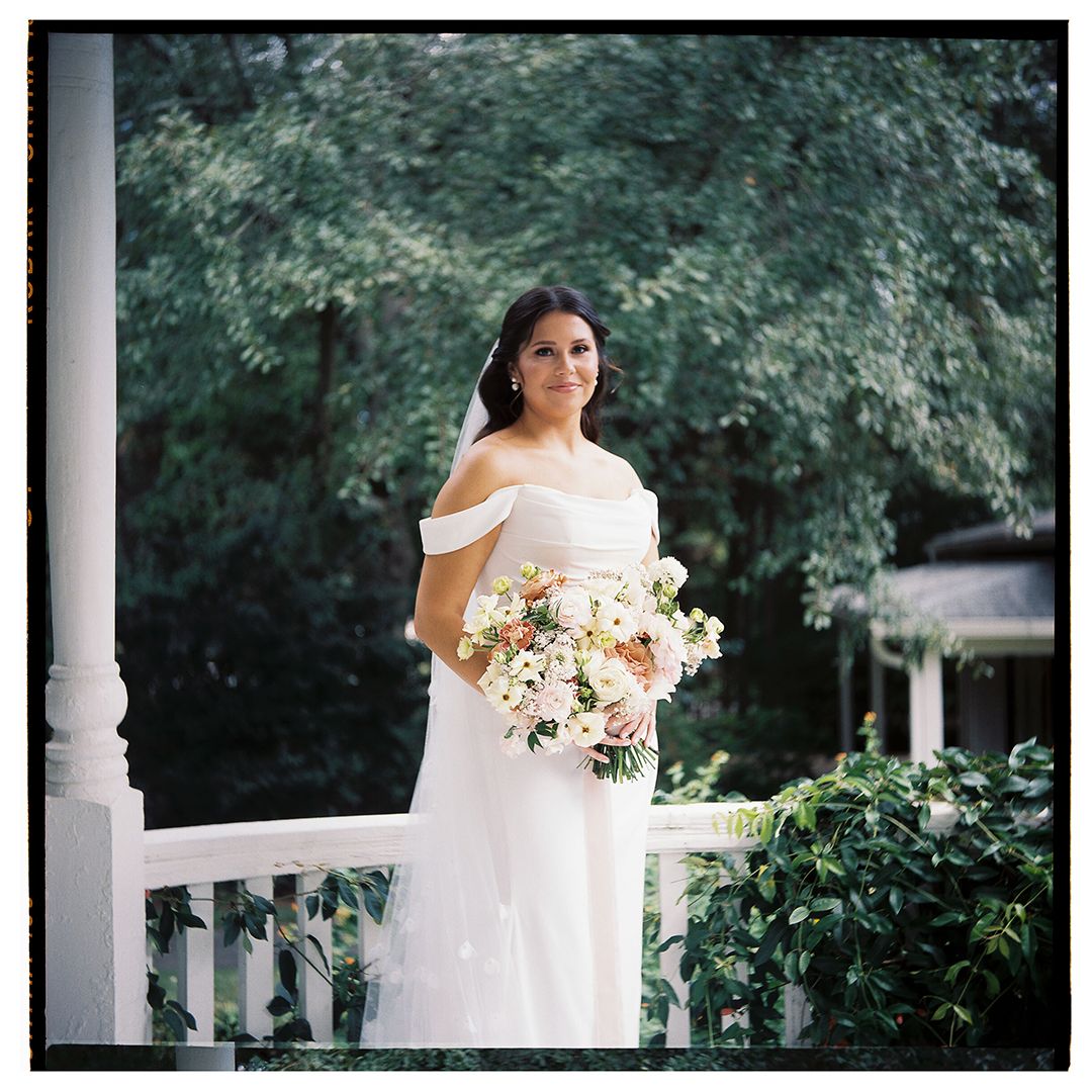 Bride in white dress holds bouquet while standing on classic white porch with columns.