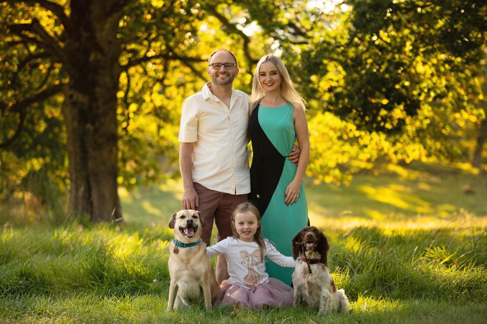 A family poses outdoors in a sunlit park setting with their three dogs during golden hour.