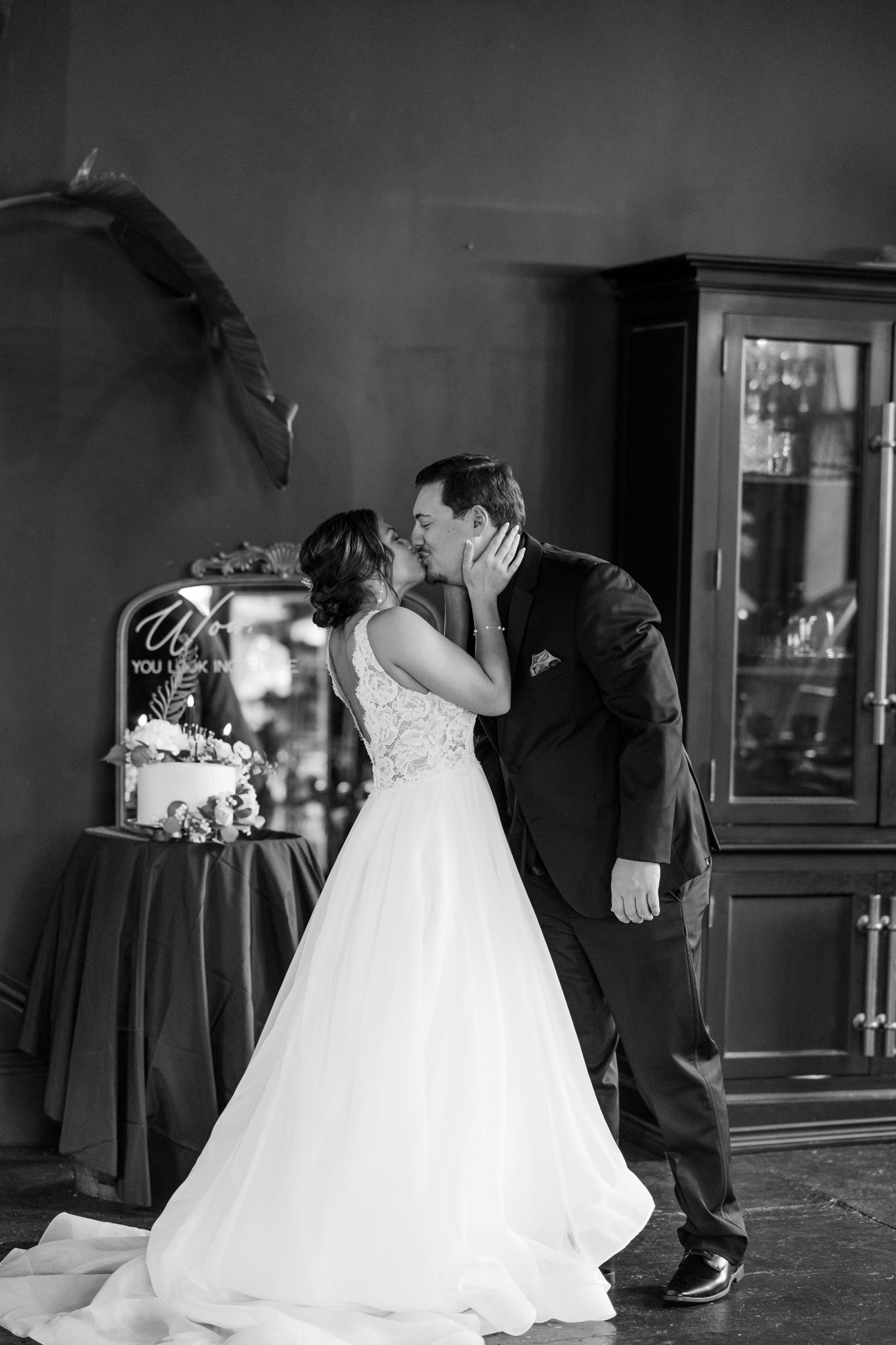 A romantic black and white photo of newlyweds sharing their first kiss by their wedding cake.