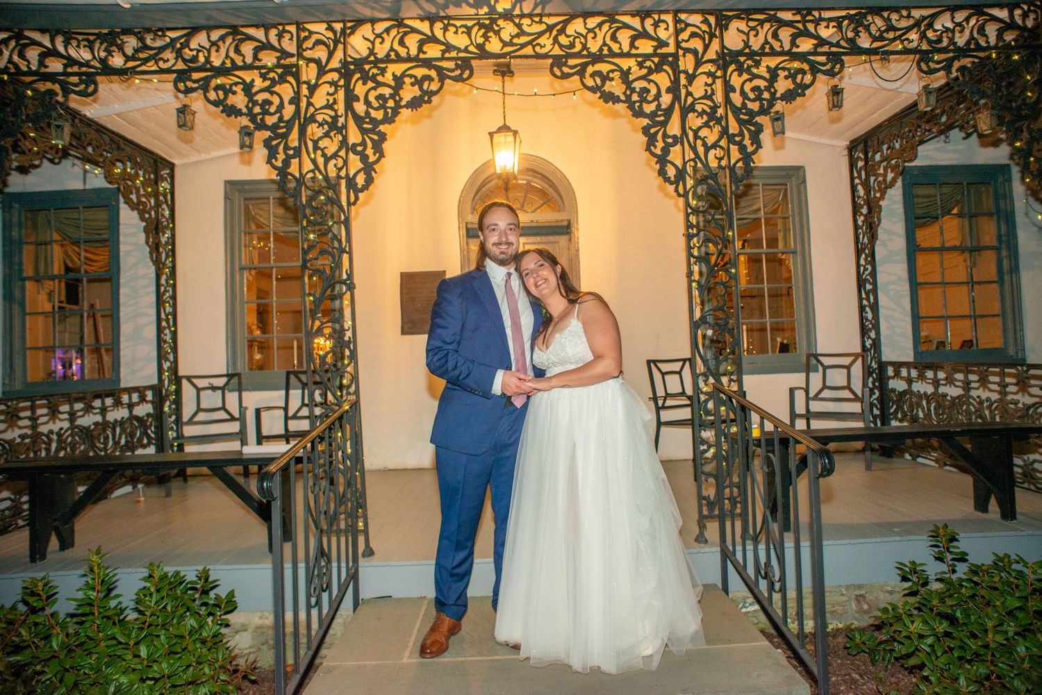 Couple poses on elegant stone staircase with ornate wrought iron railings at historic venue during sunset.