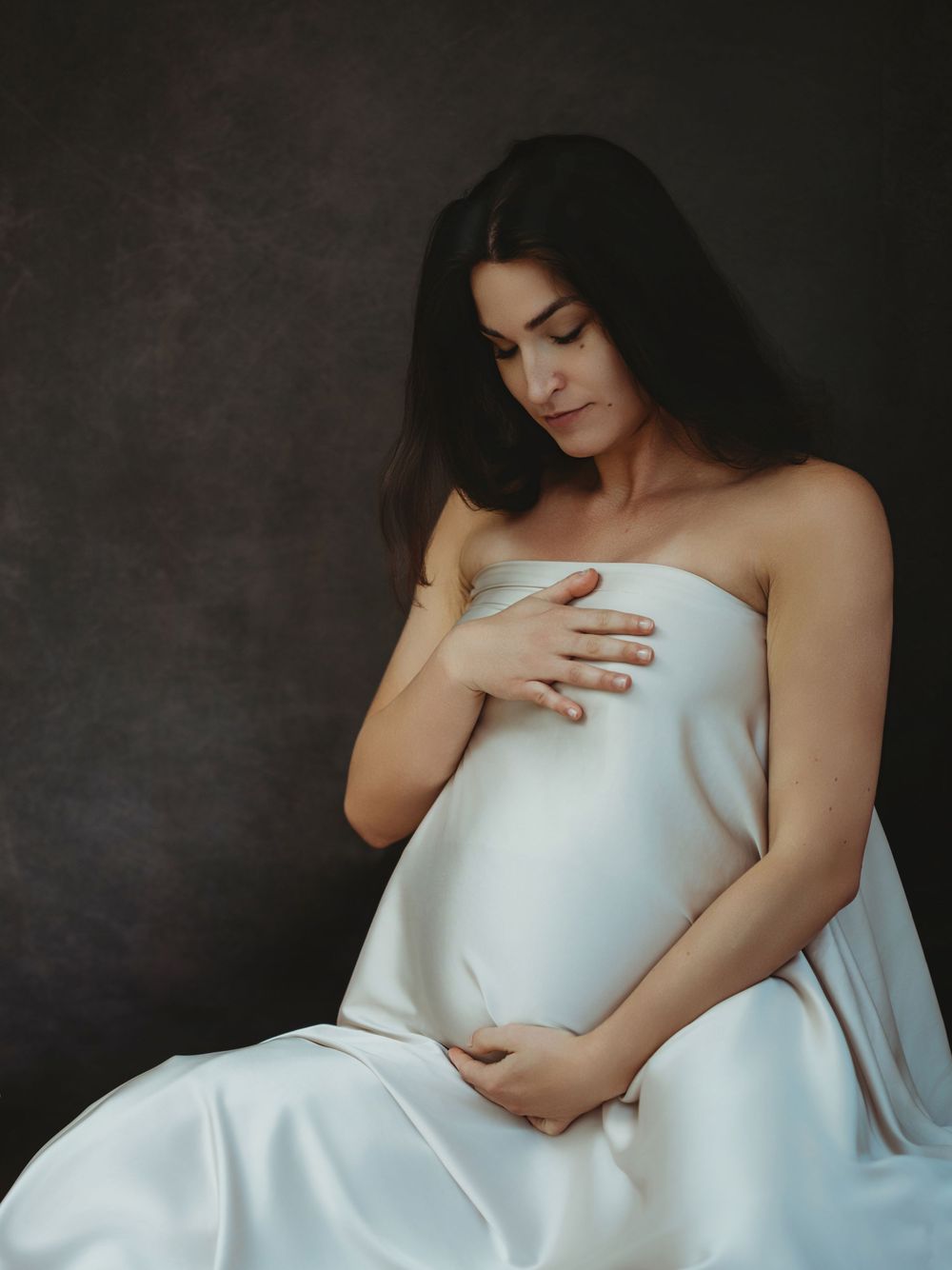 A series of artistic portraits showing a figure draped in white fabric against a dark textured background - maternity photography Bellevue