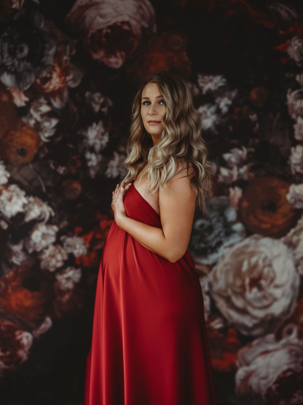 Maternity portrait against dark floral backdrop features elegant red gown and soft waves in dramatic lighting - photography Bellevue WA