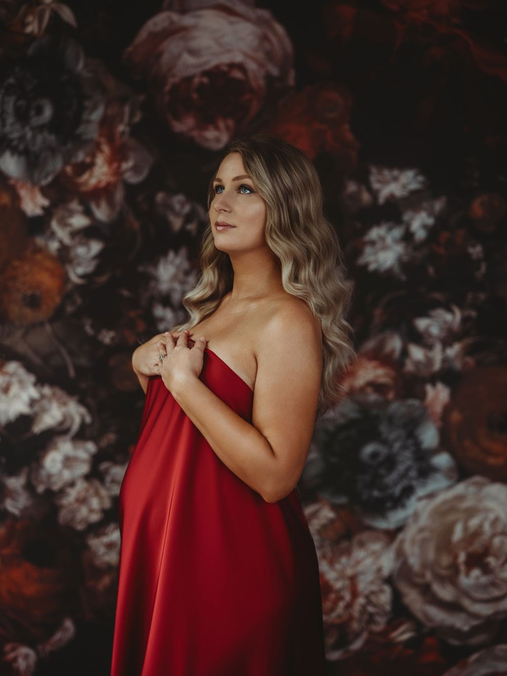 Woman in red satin dress poses against dark floral backdrop in dramatic portrait series - maternity photography Bellevue WA