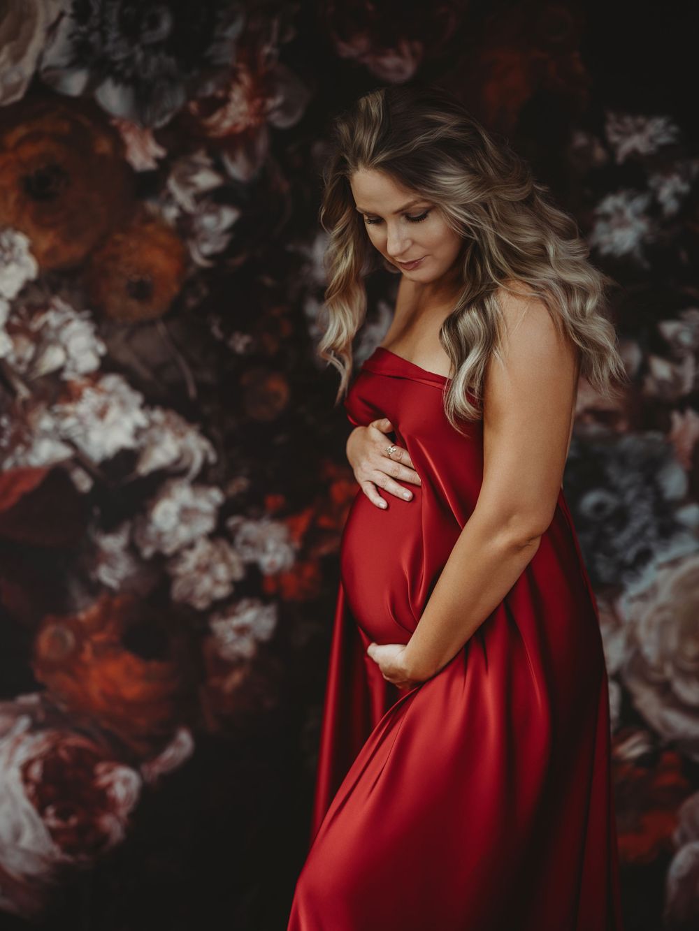 Expectant woman in flowing red dress poses against dark floral backdrop in moody maternity photoshoot - maternity photography Bellevue WA
