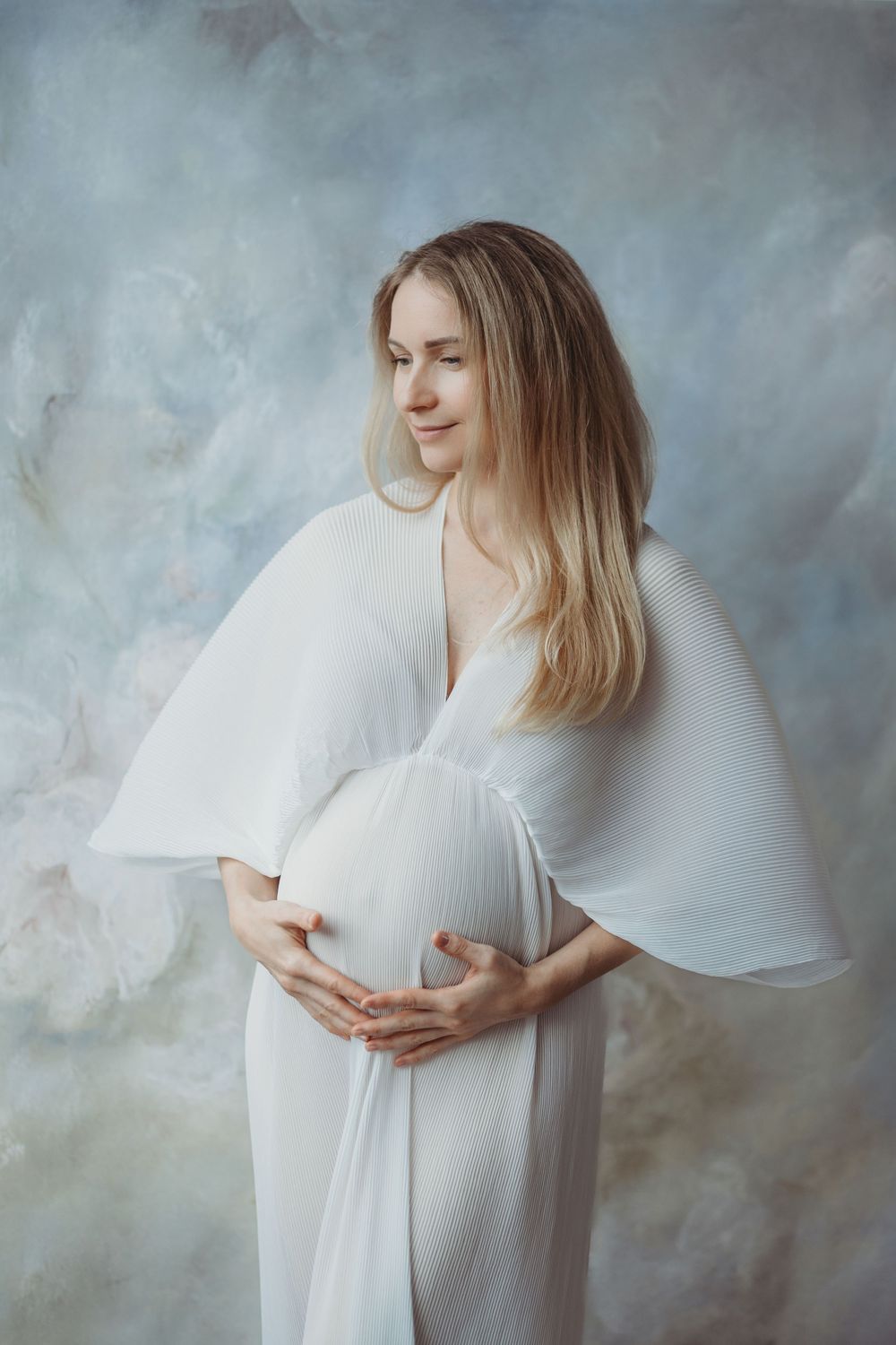 A series of ethereal maternity portraits in white against a textured gray backdrop shows gentle poses and flowing fabric - Bellevue WA
