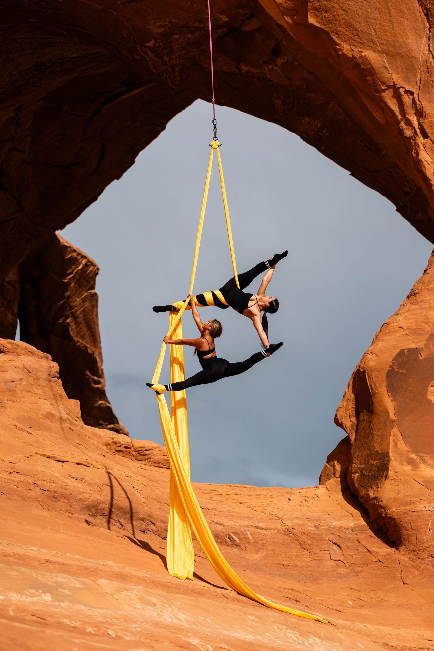 Aerialist duo on silks soaring through a Utah red rock arch; dramatic aerial arts photo merging landscape, movement, and clean form.