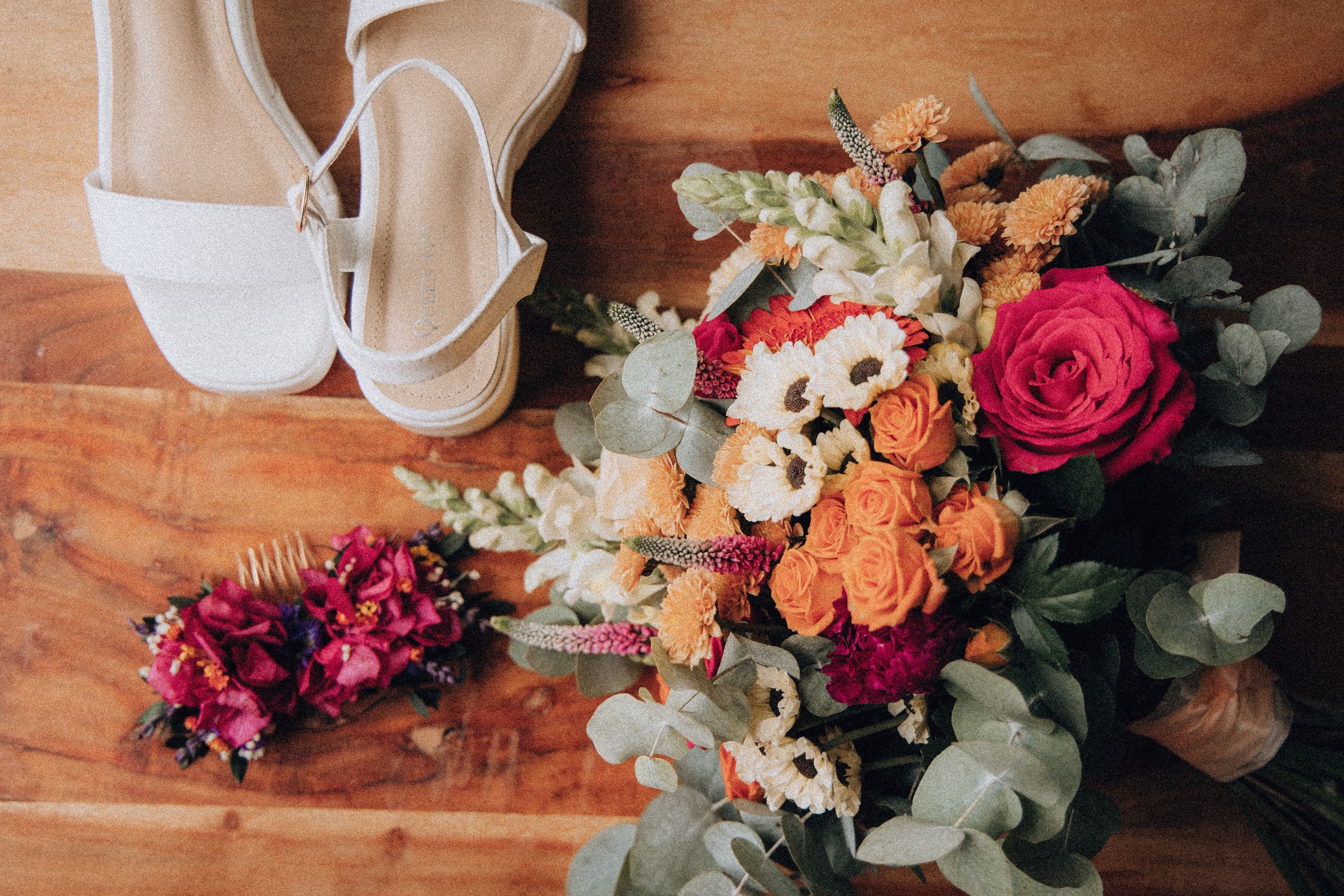 Wedding flat lay with bridal bouquet, white heels, and floral hair accessories on wood.