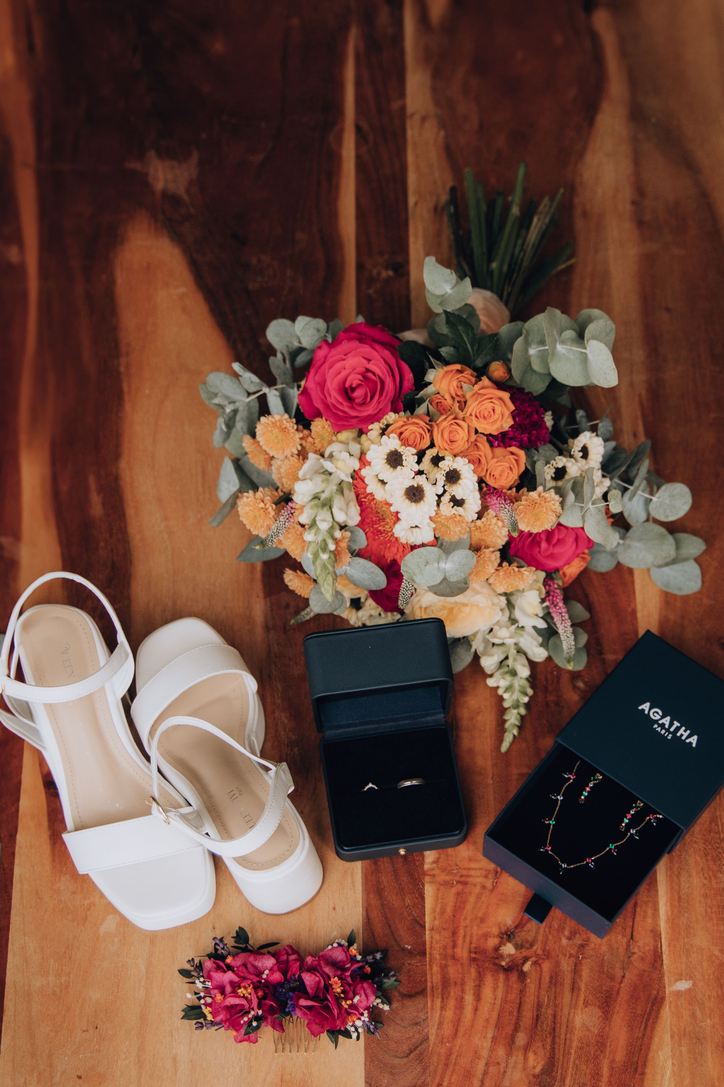 Wedding flat lay featuring bouquet, white heels, rings, and jewelry on wooden surface.