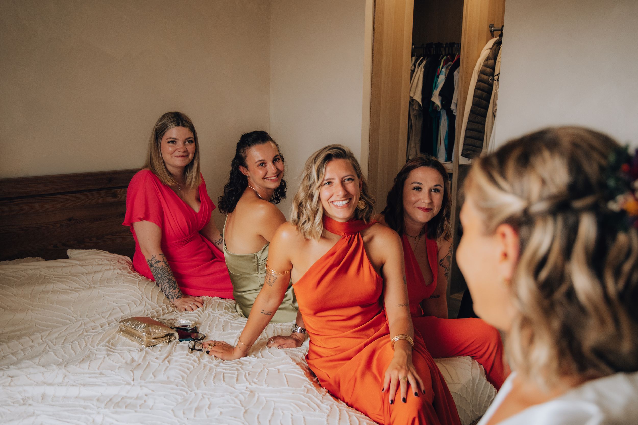 Bride smiling with bridesmaids in colorful dresses sitting together on a bed.