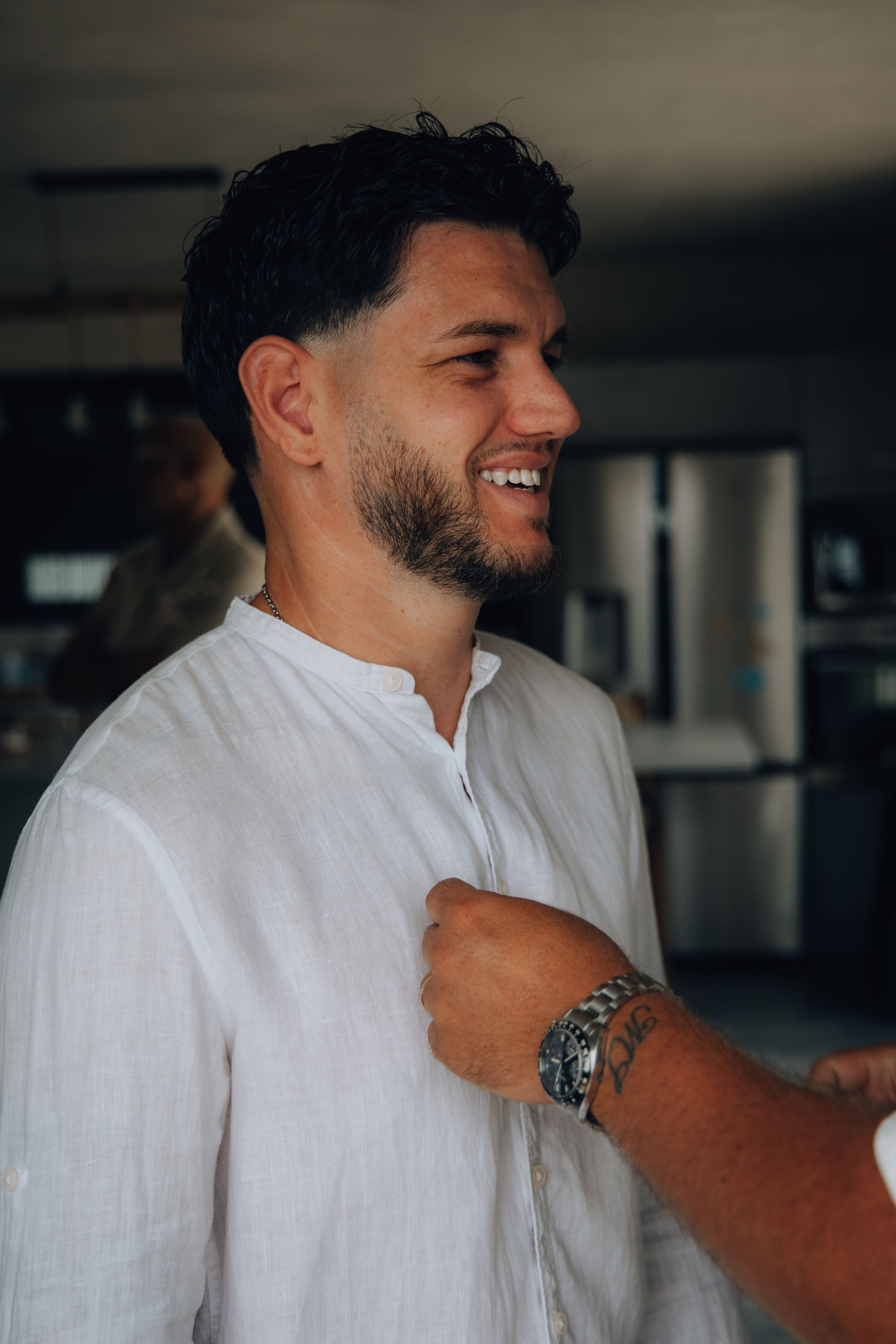 Smiling groom in a white linen shirt being helped to dress on wedding day.