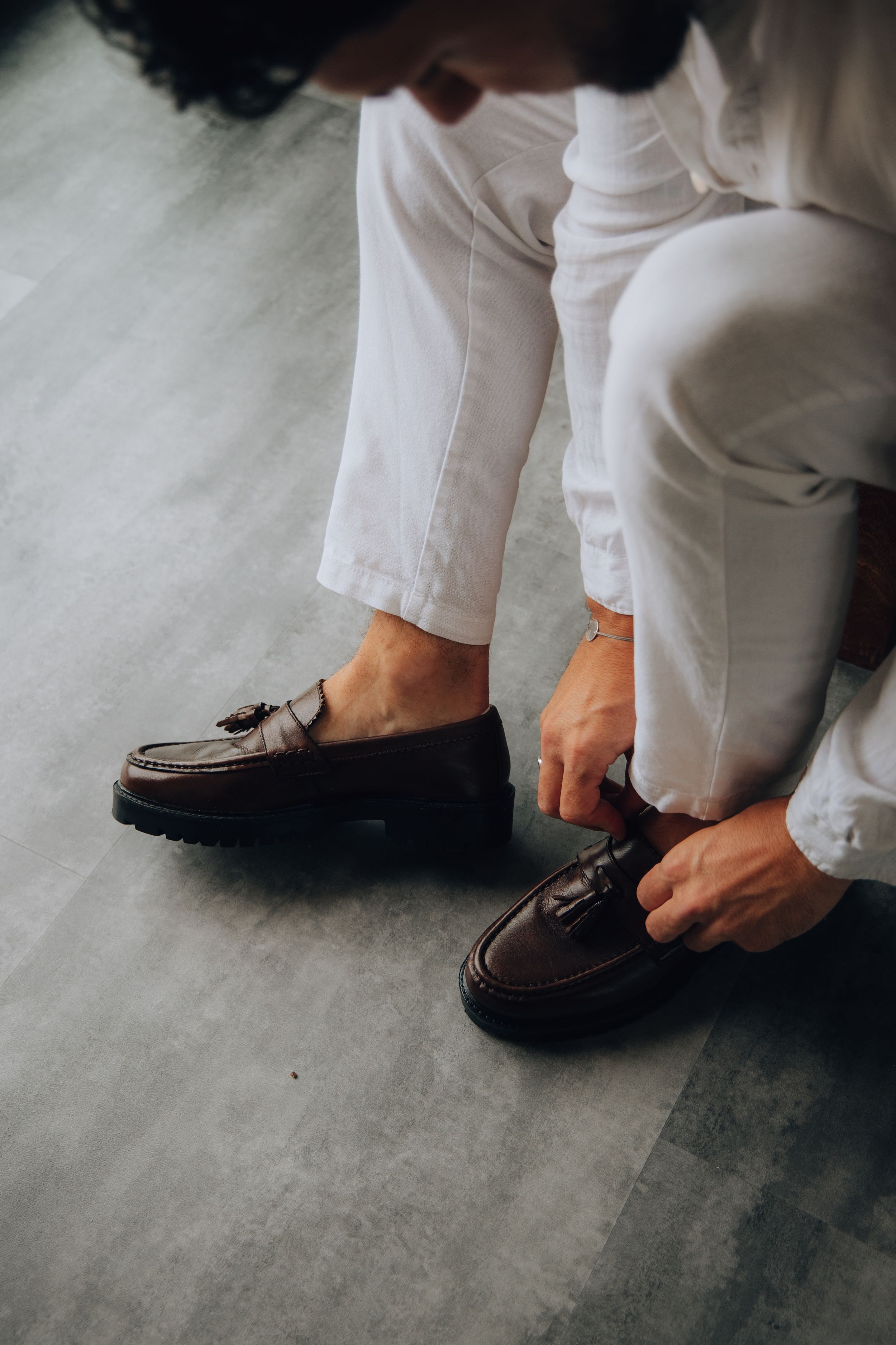 Groom in white linen trousers putting on dark brown tassel loafers on a grey floor.
