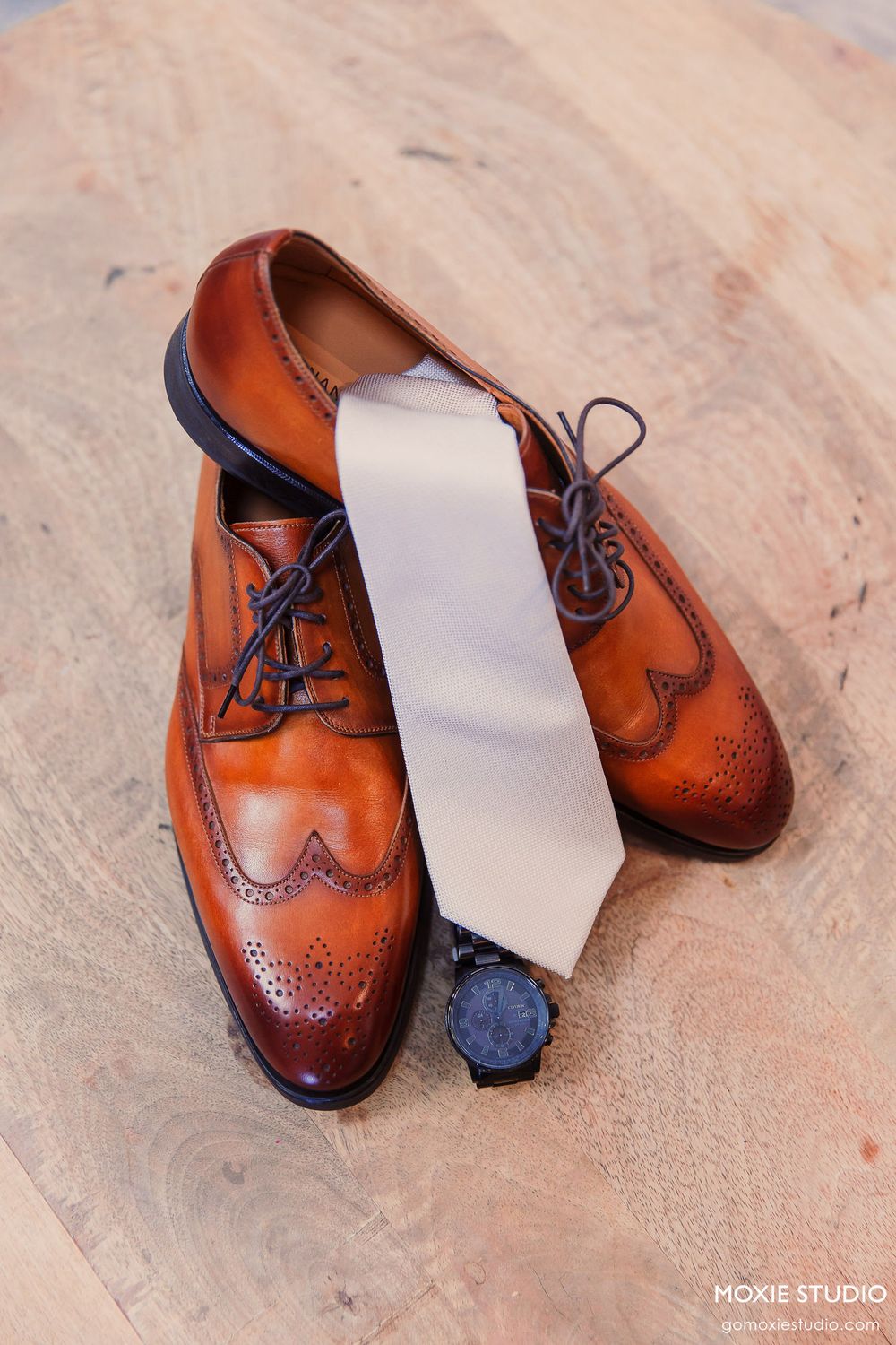 Brown leather brogue dress shoes with blue laces and a light gray necktie arranged on a wooden surface.