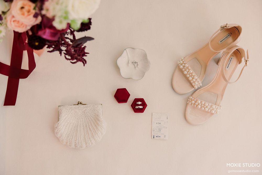 Wedding details flatlay with pearl sandals, seashell decor, burgundy flowers and delicate accessories on cream background.