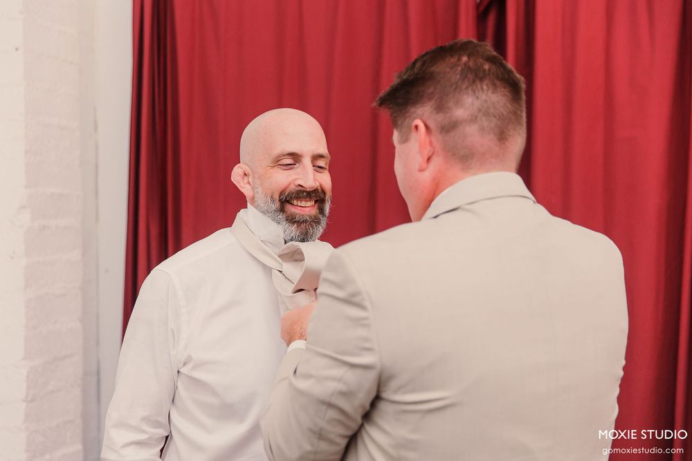 Two people in formal attire embrace warmly during a wedding ceremony against a red curtain backdrop.