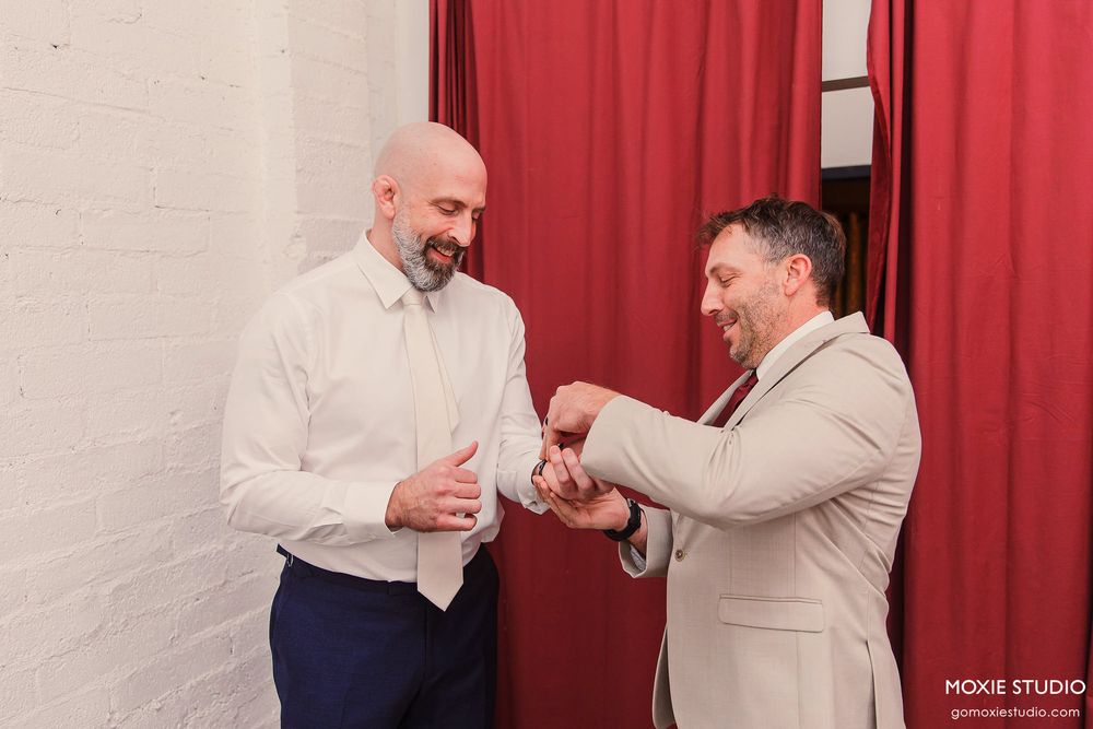 Two men in formal attire share a lighthearted moment against a backdrop of red curtains.