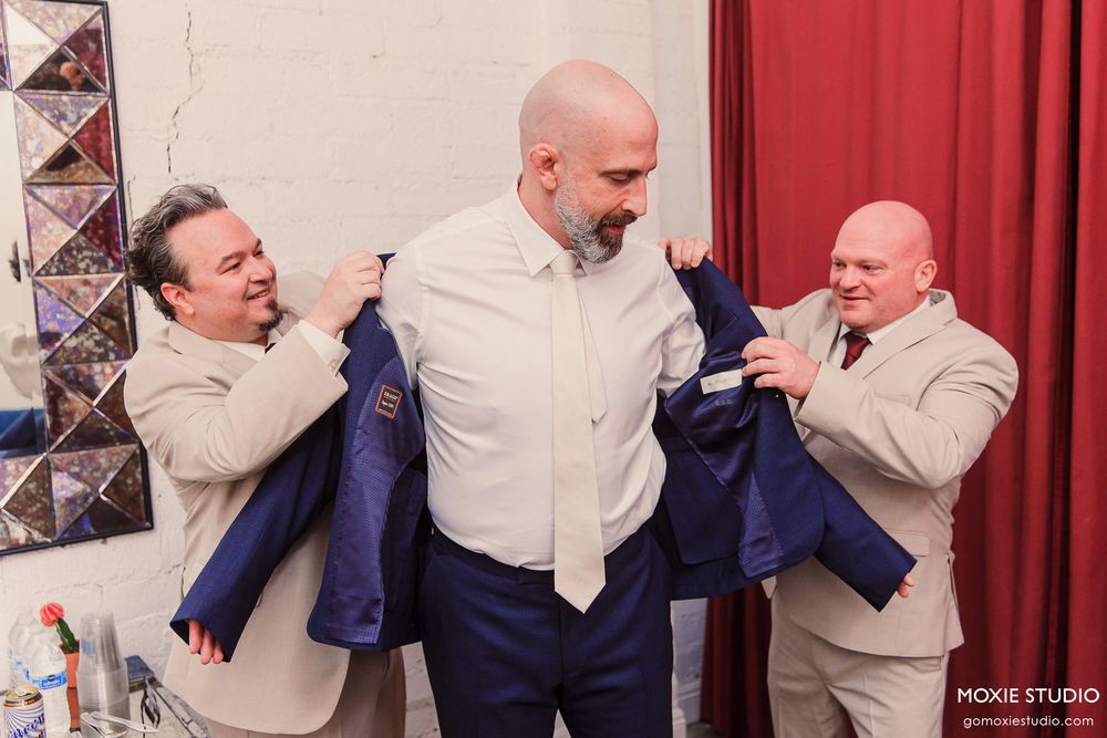 Two men help another man put on a blue suit jacket while getting ready in front of red curtains.