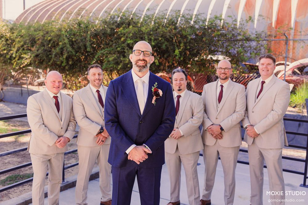 Wedding party members in light beige suits and navy blue attire pose together outdoors for formal group photos.
