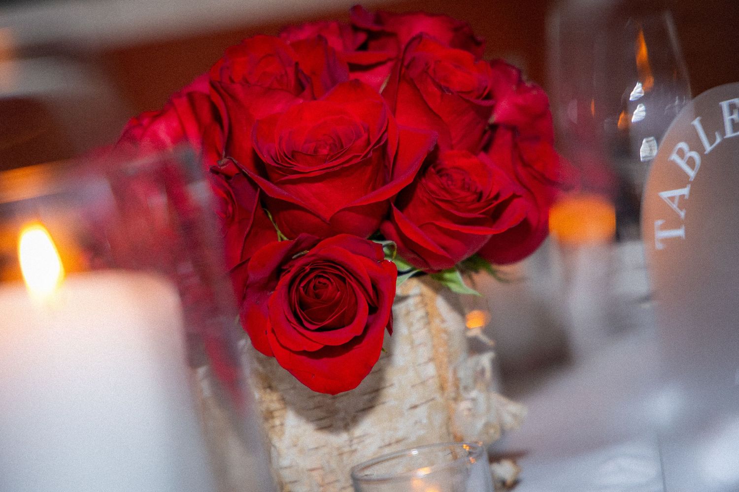 A romantic centerpiece of vibrant red roses in a rustic vase with a glowing candle in soft focus.