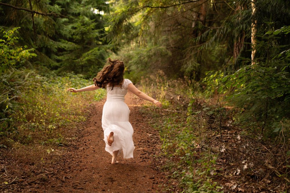 A musician in a white dress plays acoustic guitar in a lush green forest setting during golden hour. Captured by Savvy Photography.