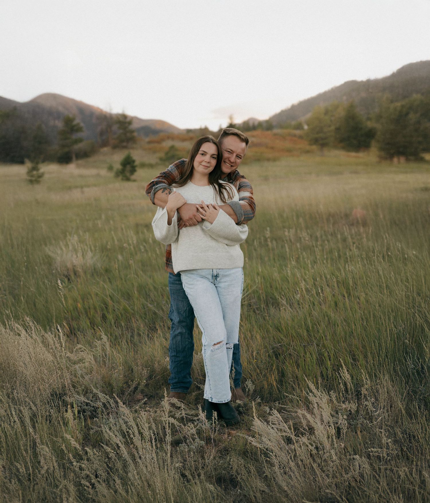 Couple embraces in a golden field with mountains in the background during sunset creating a romantic outdoor scene.