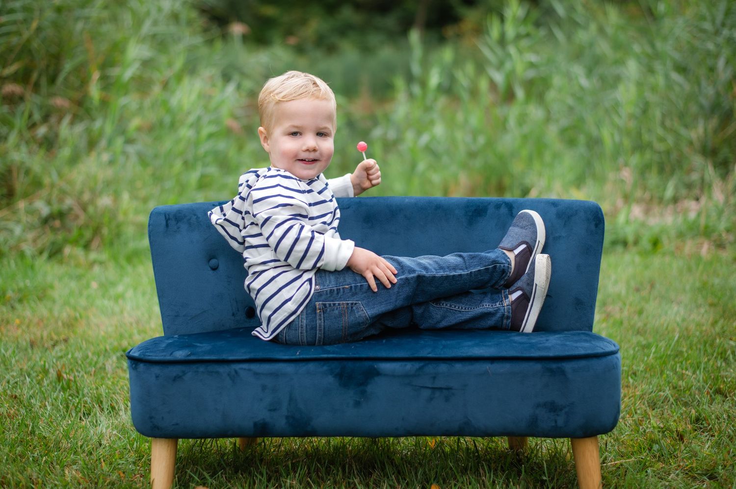 A toddler in striped clothing plays and poses on a navy blue couch outdoors in a grassy setting.
