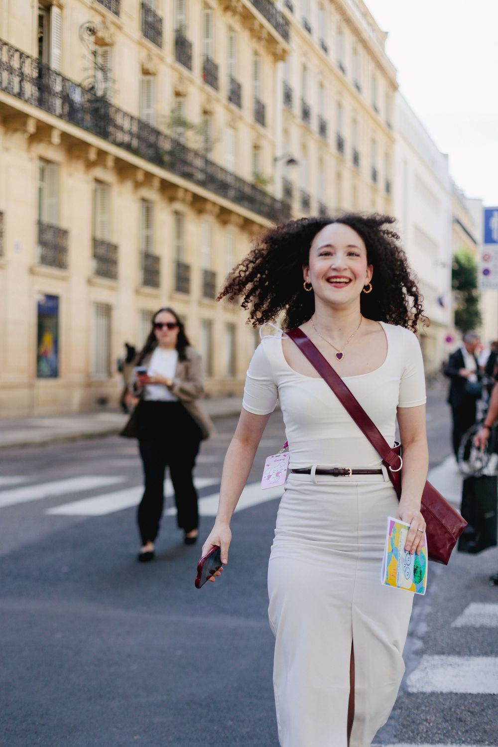 Smiling curly-haired woman in beige outfit crossing a Parisian street on a sunny day.
