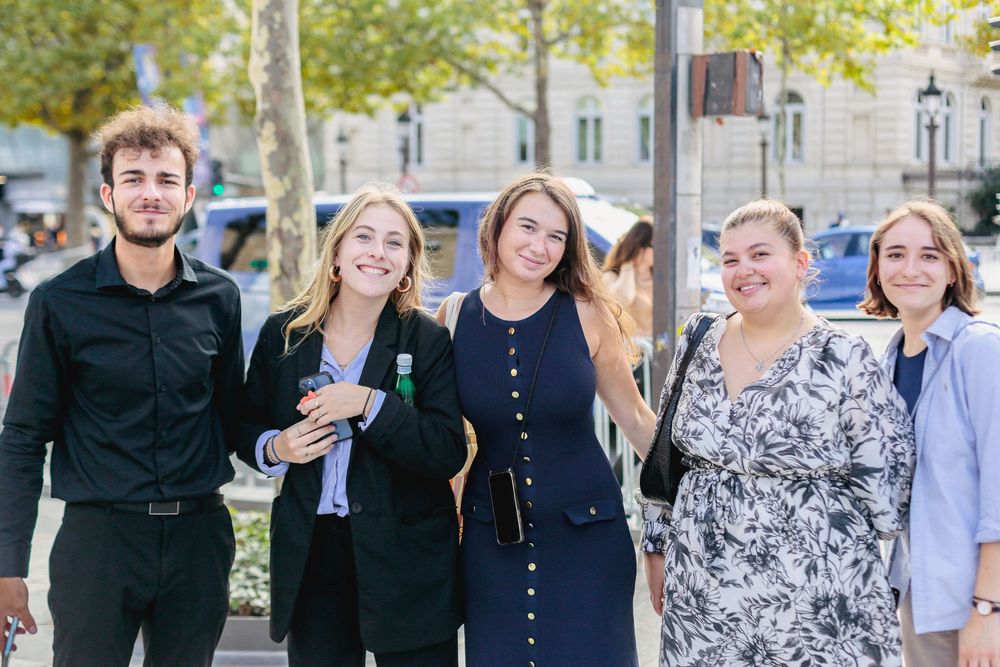 Five young students smiling together on a sunny tree-lined Paris street.