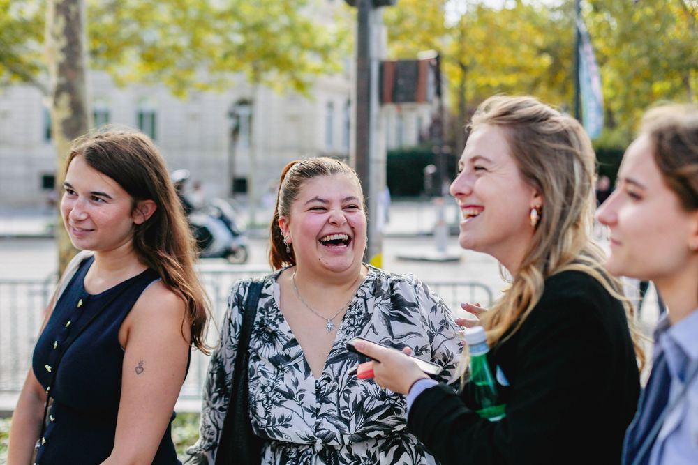 Four young women laughing together outdoors at a sunny Paris gathering.