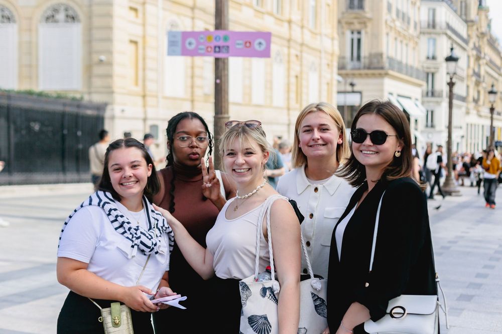 Five diverse young women posing together on a sunny Paris shopping street.