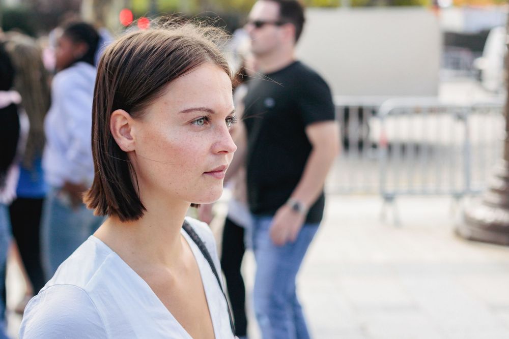 Young woman with short brown hair in white top standing outdoors in crowd.