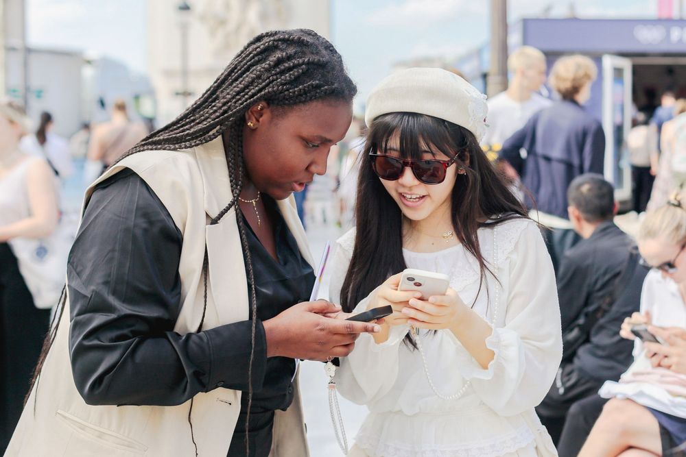 Two stylish women sharing phones at a sunny outdoor Paris event.