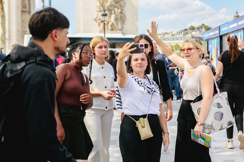 Group of young people taking selfies in a sunny Paris public square.