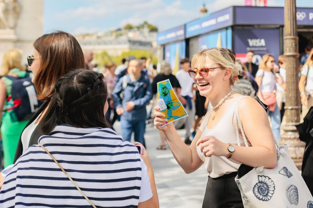 Smiling woman photographing friends with smartphone at sunny Paris outdoor event.