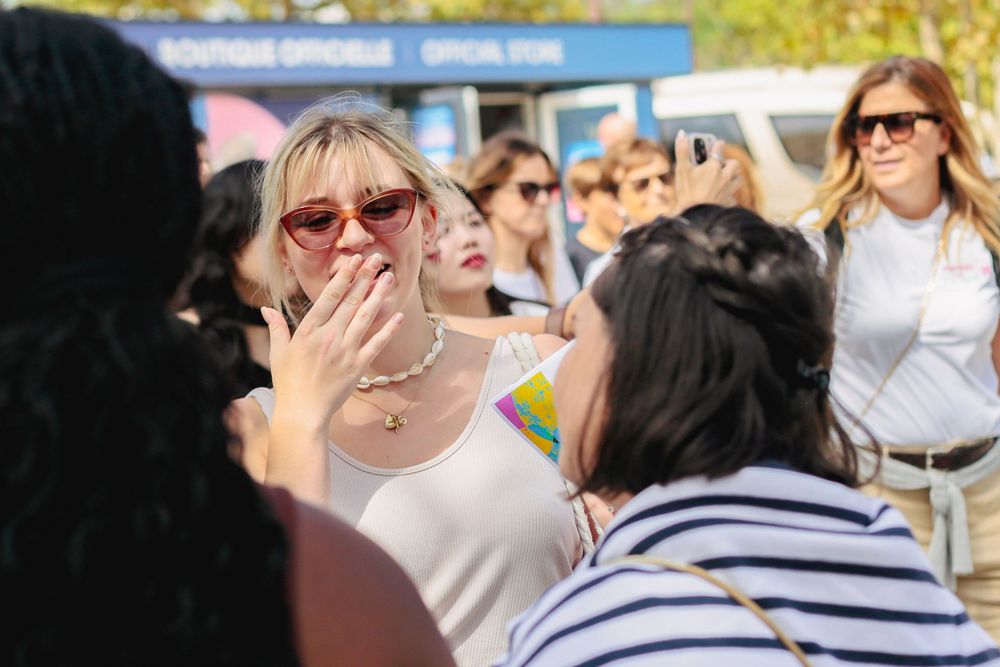 Emotional woman in sunglasses covers her mouth in surprise while surrounded by fans outdoors.