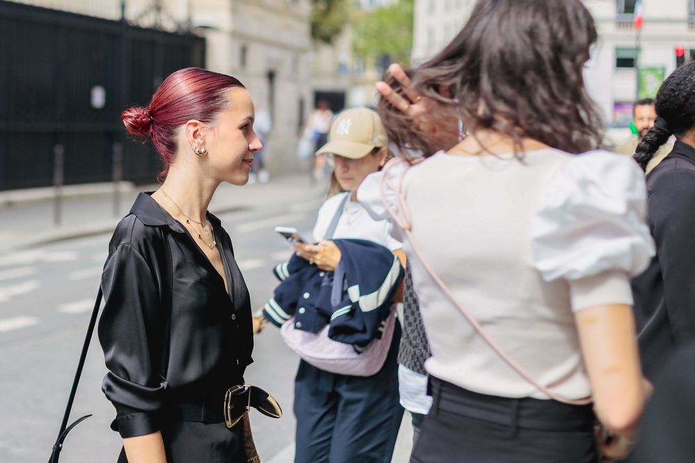 Two stylish women with colorful hair chat on a busy urban street in daylight.
