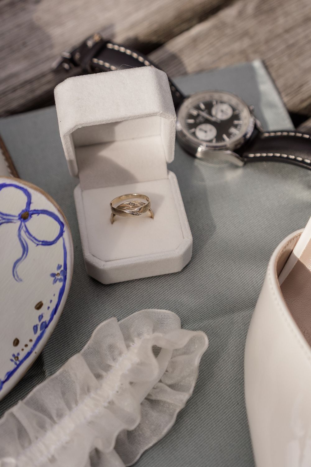 Wedding detail photo showing white ring boxes, a delicate lace garter, and blue decorative accents on a gray surface.