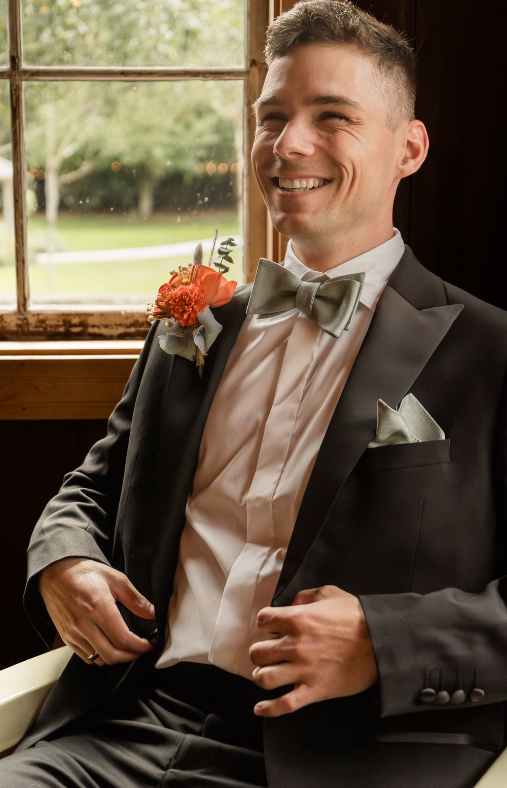 Groomsman in black tuxedo and bow tie smiles while sitting in vintage car at wedding.