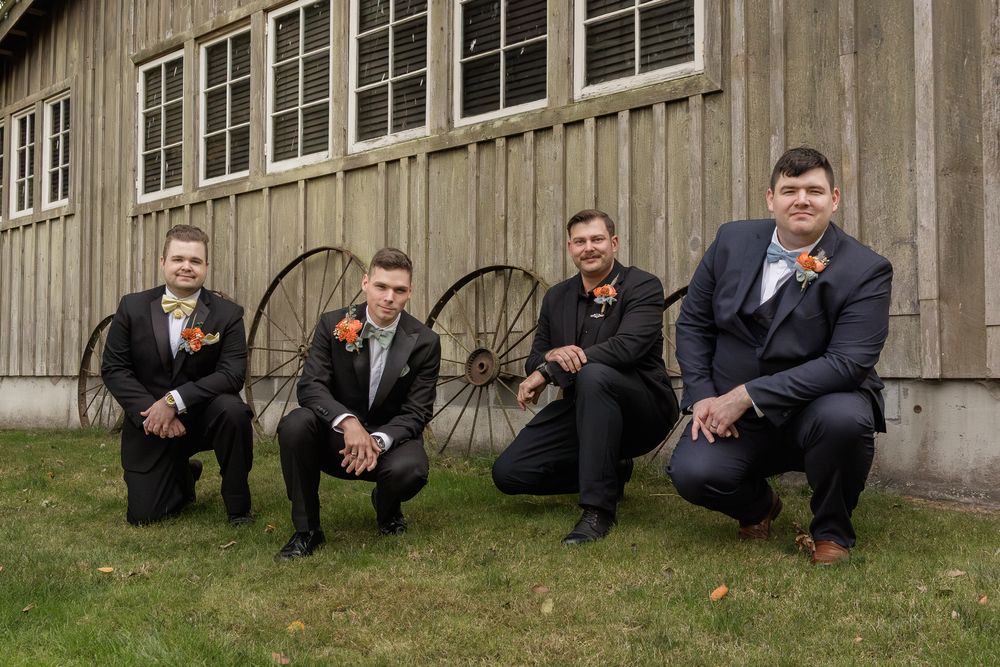 Groomsmen in dark suits crouch in line against rustic wooden barn wall during wedding photo session.