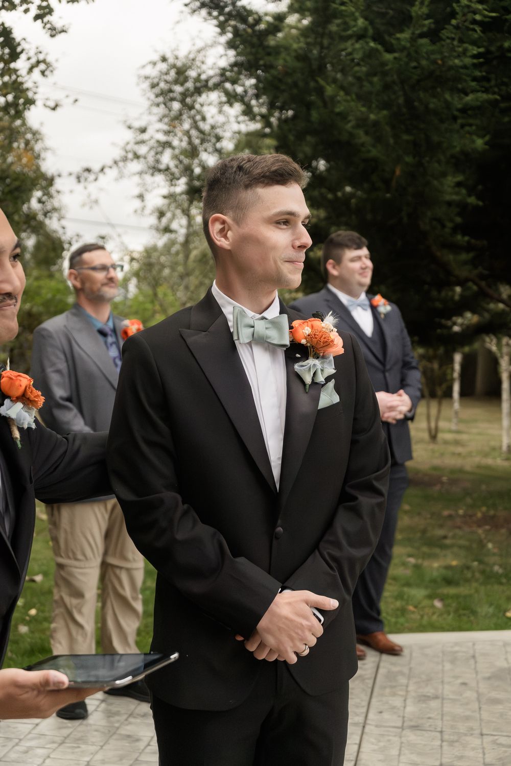 Groomsmen in dark suits and orange boutonnieres stand in a line outdoors during a wedding ceremony.